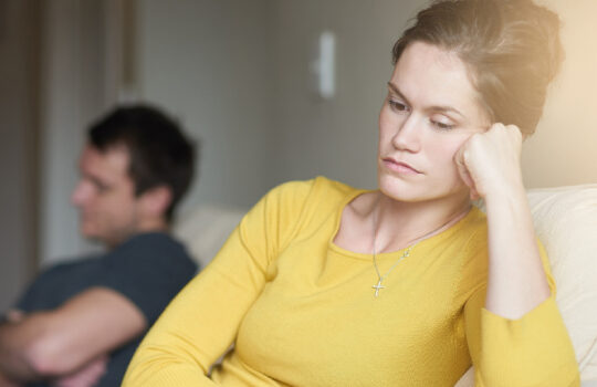 a couple sitting apart on a couch looking upset